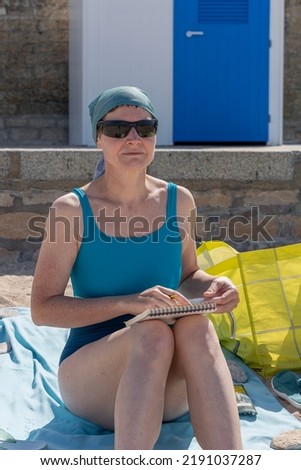 A woman sitting on the sand of the beach wearing sunglasses, a blue cap and a blue swimsuit is drawing