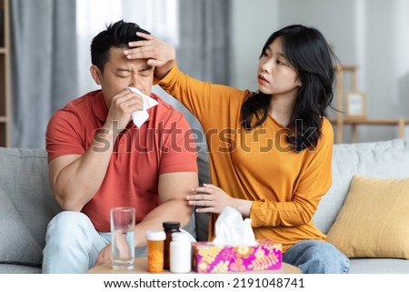 Young asian wife taking care of sick husband sitting on sofa next to tea table full of medicine and sneezing nose, loving woman touching her ill boyfriend forehead. Cold, flu, coronavirus concept Royalty-Free Stock Photo #2191048741
