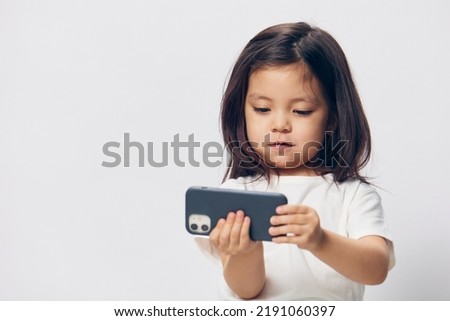  little girl in a white t-shirt stands on a light background with a fashionable smartphone in her hand and takes a selfie, looking at the phone. photography with empty space for advertising mockup