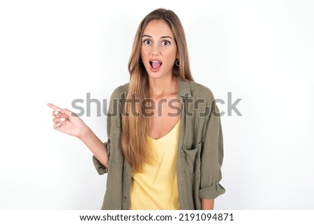 Emotive young beautiful woman wearing green overshirt over white background keeps jaw dropped from shock demonstrates amazing promo points right on blank space demonstrates big shopping sale. 