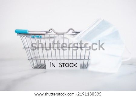 In stock text in front of empty shopping basket on white background with surgical mask, concept of supply chain shortages and delays after the covid-19 global pandemic