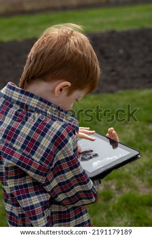 Happy red-haired boy playing on a tablet or watching cartoons, walking in the backyard in the village, summer holidays, view from the back.