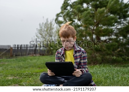 Happy redhead boy playing tablet or watching cartoons sitting on green grass in the backyard in the village.