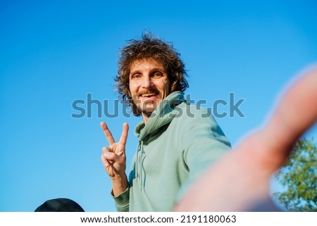 A happy guy takes a selfie, a self-portrait on his phone against the sky, a curly man smiling, hand gestures, a cheerful hipster. High quality photo
