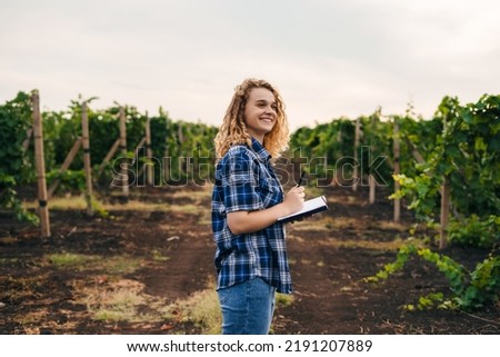 Smiling curly woman examines the vineyard and recording data in the notebook. Farmer field. Data analysis. Royalty-Free Stock Photo #2191207889