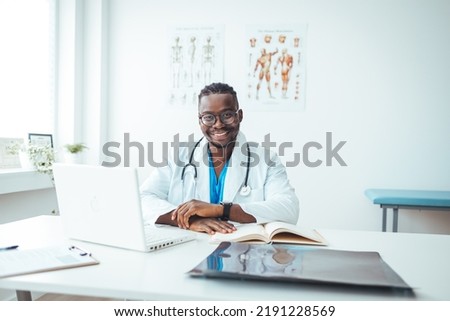 Portrait Of Smiling male Doctor Wearing White Coat With Stethoscope Sitting Behind Desk In Office. Happy male medical doctor portrait in hospital. Portrait of a male doctor with laptop sitting at desk