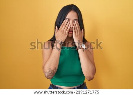Brunette woman standing over yellow background rubbing eyes for fatigue and headache, sleepy and tired expression. vision problem 
