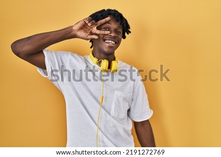 Young african man with dreadlocks standing over yellow background doing peace symbol with fingers over face, smiling cheerful showing victory 