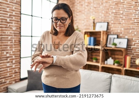 Middle age hispanic woman looking smartwatch standing at home