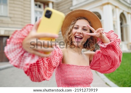 Young smiling woman in pink dress hat doing selfie shot on mobile cell phone post photo on social network show v-sign gesture walk in city center stand outdoor near building Urban lifestyle concept