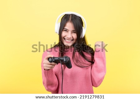 Beautiful Asian woman playing games and wearing wireless headphones standing on isolated yellow background. Play  game on your smartphone that wins the moment of victory. Fun and enjoyable vacation 