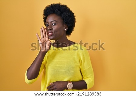 African young woman standing over yellow studio hand on mouth telling secret rumor, whispering malicious talk conversation  Royalty-Free Stock Photo #2191289583
