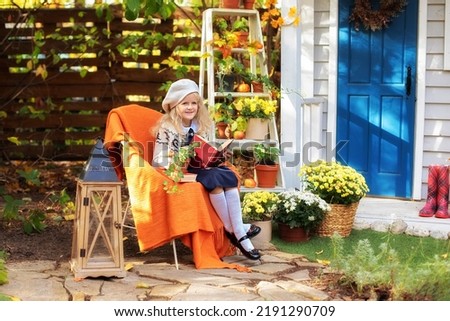 Little smiling girl sit on chair and reading book in fall garden. Child girl rest with book sitting porch home with fall decor and flowers. Kid is sit next to pumpkins and flowers pots near of house.