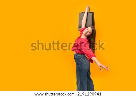 Cheerful young girl with black paper bags in her hands after successful shopping on yellow background