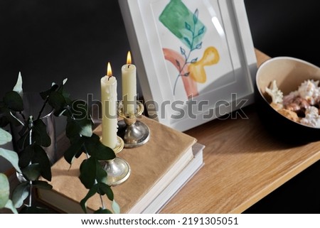 interior and home decor concept - close up of bench with burning candles, picture in frame, books, seashells and eucalyptus branches over black background