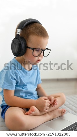 A child in glasses and headphones sits cross-legged and looks into a laptop. 