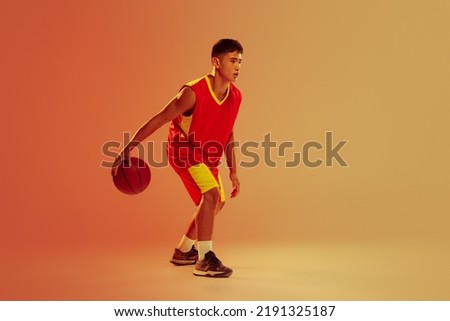 Portrait of young man, basketball player training isolated over orange studio background in neon light. Dribbling exercises. Concept of healthy lifestyle, professional sport, hobby, power and