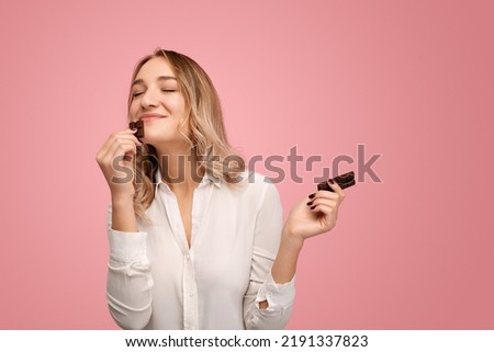 Content young female with long blond hair in white shirt smiling while enjoying smell of delicious chocolate with closed eyes against pink background Royalty-Free Stock Photo #2191337823