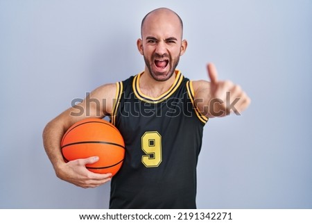 Young bald man with beard wearing basketball uniform holding ball approving doing positive gesture with hand, thumbs up smiling and happy for success. winner gesture. 