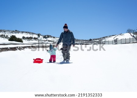 Skier father and his little daughter walking on an empty ski slope with a red sledge. Winter activities in family.