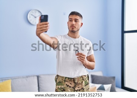 Young hispanic man wearing camouflage army uniform taking selfie at home relaxed with serious expression on face. simple and natural looking at the camera. 