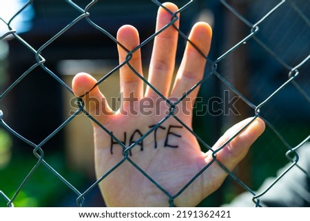 Caucasian man behind wired fence showing hes palm with the word hate, focus on the wired fence.