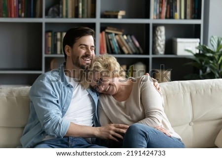 Overjoyed mature senior retired woman leaning on shoulder of happy grown up son, laughing at funny story together while relaxing on comfortable couch in modern living room, weekend family pastime.