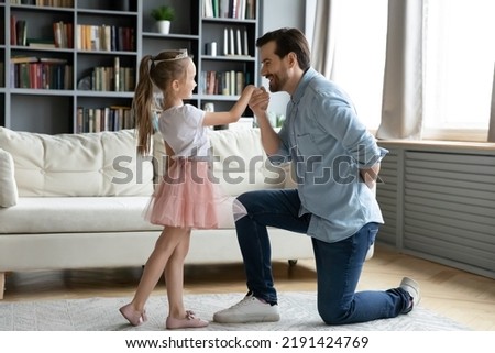 Full length gentle loving caring young father standing on knee, kissing hand of little princess dressed adorable child daughter, inviting for dance in living room, domestic hobby playtime activity.