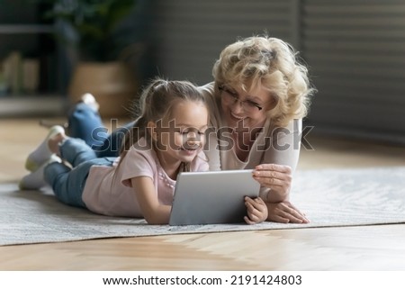 Full length cute happy little preschool child girl showing funny application to affectionate middle aged 60s grandmother, lying together on floor carpet in living room, technology addiction concept.