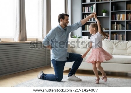 Joyful young father standing on one knee, raising arms twisting little funny preschool kid daughter, dressed as princess in living room. Happy different generations family dancing to music at home. Royalty-Free Stock Photo #2191424809