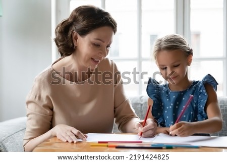 Close up smiling mature grandmother and granddaughter drawing colored pencils together, sitting at table in living room, middle aged woman babysitter teacher and child engaged in creative activity