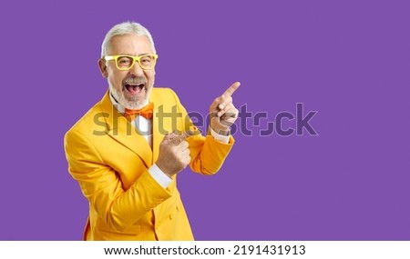 Cheerful extravagant and funny senior man pointing fingers at copy space on purple background. Gray-haired man in yellow suit and glasses with funny expression advertises or recommends. Banner.