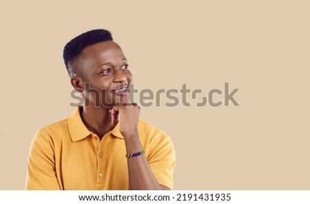 Young happy African American man student looks away with smile remembers home and loved ones props head up with hand dreams of happy future and career stands on beige studio background Royalty-Free Stock Photo #2191431935