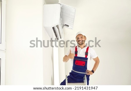 Happy young man in uniform standing on ladder under new air conditioner unit in white room, smiling and showing thumbs up after a job well done. AC installation, maintenance and repair service