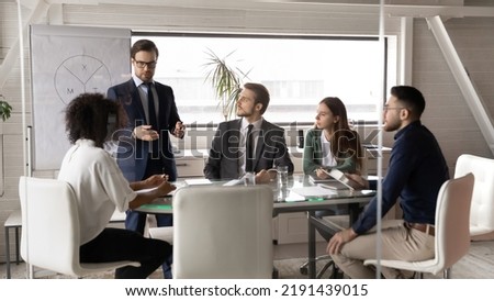 Confident young Caucasian businessman stand lead meeting with multiracial colleagues in office. Successful male coach or CEO talk make flip chart presentation at briefing with diverse businesspeople