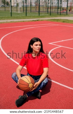 A girl holds a basketball in her hands, listens to music, resting after a game at an outdoor stadium on a hot sunny day.