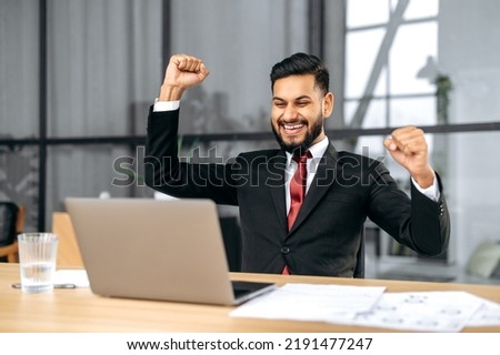 Excited indian or arabian businessman in suit, sits in a modern office, looking at laptop, happy by good news, rejoice by win, raising hand in yes gesture, celebrating project success, big profit Royalty-Free Stock Photo #2191477247