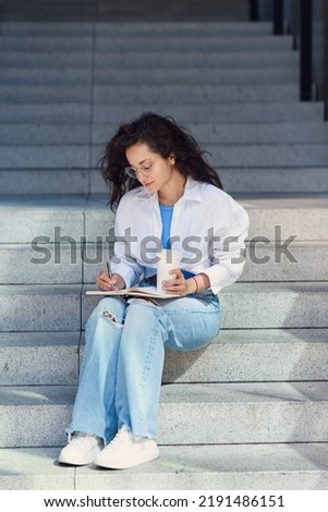 Adorable young office female worker in white shirt with book and coffee sitting on stairs outdoors.