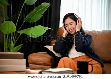 Attractive and joyful young Asian female enjoys listening to music on her wireless headphone while relaxing on the sofa in her living room.