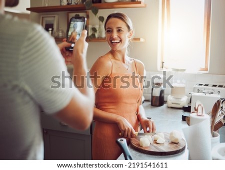 Woman cooking with husband taking a picture in kitchen on his mobile phone at home. Beautiful, happy and female chef cutting vegetables to cook healthy, delicious and vegetarian food at a table.
