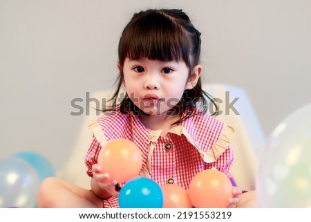 Portrait studio shot of little cute kindergarten preschooler kid girl daughter in red long dress sitting smiling alone on floor playing with colorful helium air party balloons on gray background.
