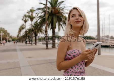 Romantic charming woman with blond hairstyle and beautiful make up is holding smartphone and looking aside with happy smile. Charming lady in wireless headphones on blur background with palm trees. 