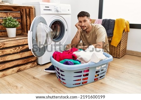 Young handsome man putting dirty laundry into washing machine thinking looking tired and bored with depression problems with crossed arms. 