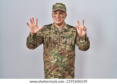 Hispanic young man wearing camouflage army uniform showing and pointing up with fingers number eight while smiling confident and happy. 
