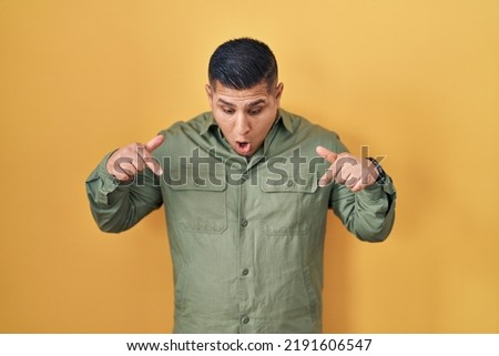 Hispanic young man standing over yellow background pointing down with fingers showing advertisement, surprised face and open mouth 
