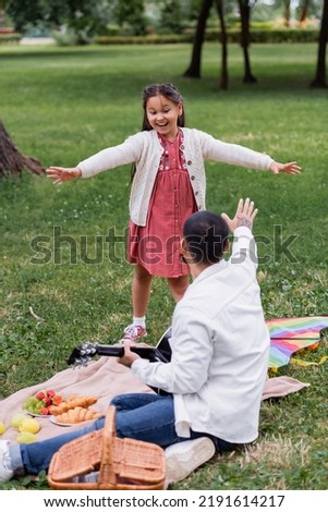 Positive asian girl looking at dad holding acoustic guitar near food on blanket in park