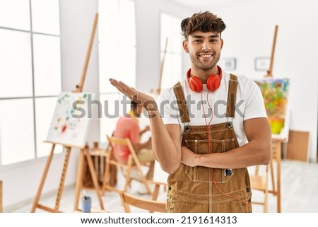 Young hispanic man at art studio smiling cheerful presenting and pointing with palm of hand looking at the camera. 