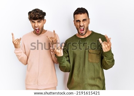 Young gay couple standing over isolated white background shouting  doing rock symbol with hands up. music star. heavy concept. 