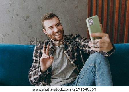 Young happy man 20s he wears brown shirt hold in hand use mobile cell phone talk v-sign sitting on blue sofa in own living room apartment stay home indoor flat on weekends People lifestyle concept.