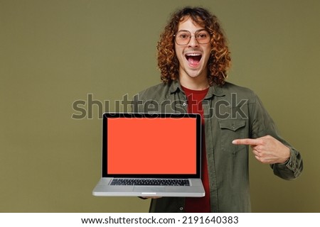Excited young brunet curly man 20s wears khaki shirt hold use work pointing index finger on laptop pc computer with blank screen workspace area isolated on plain olive green background studio portrait
